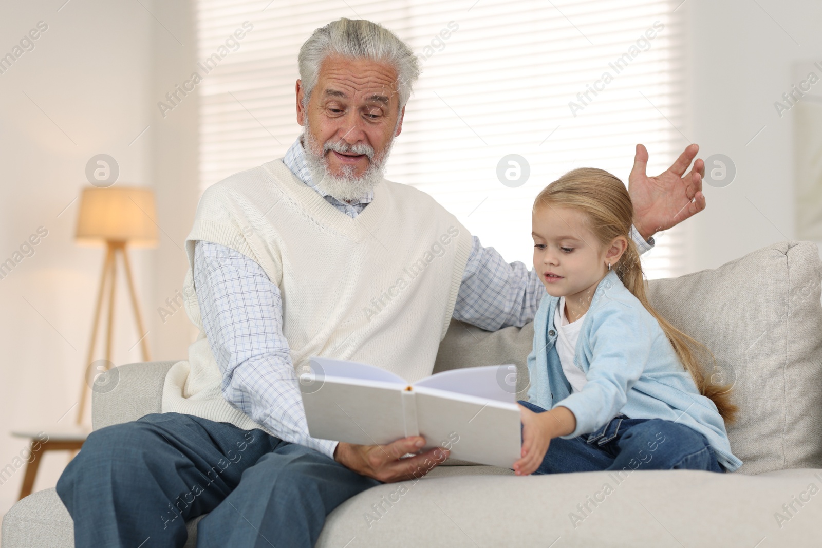 Photo of Grandpa and his granddaughter reading book together on sofa at home