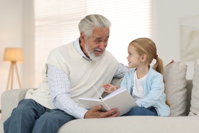 Photo of Grandpa and his granddaughter reading book together on sofa at home