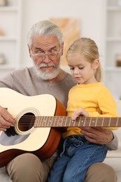 Grandpa teaching his granddaughter to play guitar on sofa at home