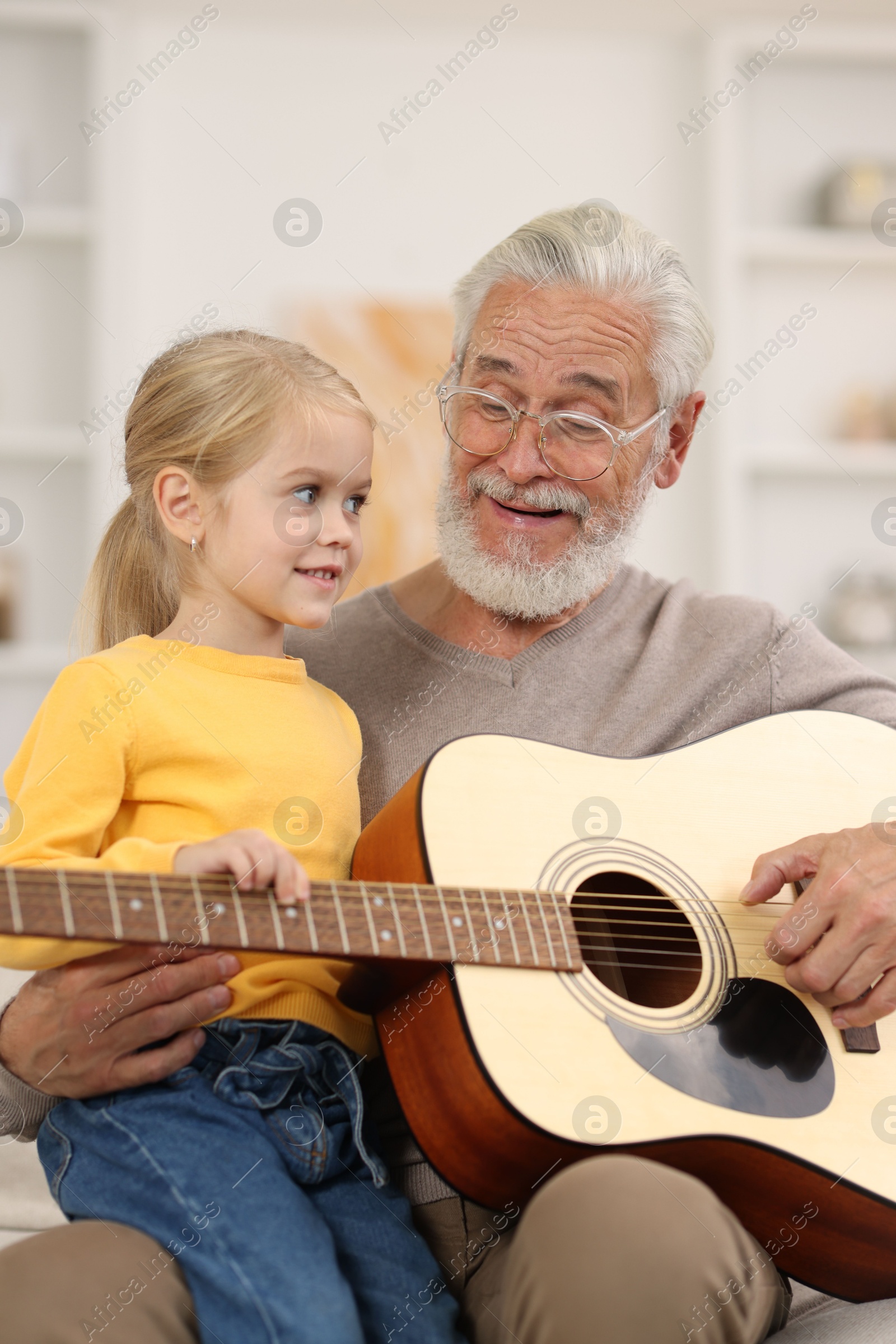 Photo of Grandpa teaching his granddaughter to play guitar on sofa at home