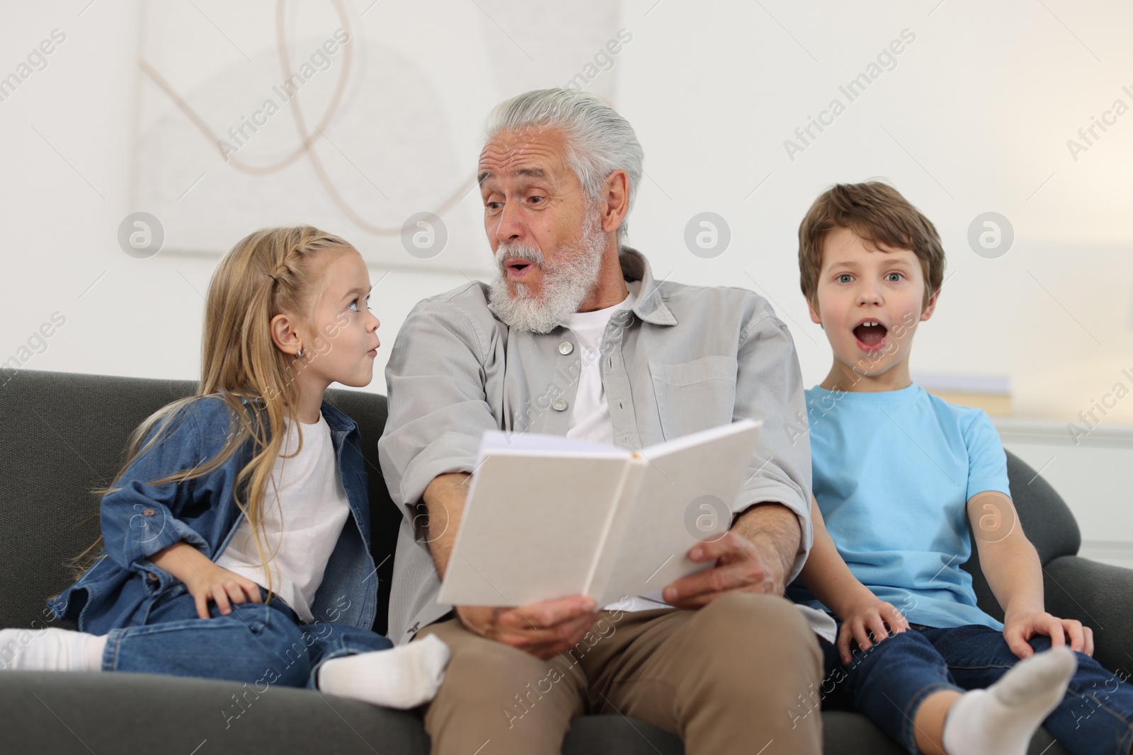 Photo of Grandpa and his grandkids reading book together on sofa at home