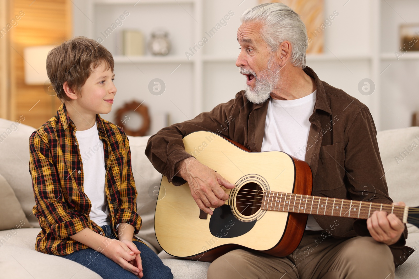 Photo of Grandpa teaching his grandson to play guitar on sofa at home