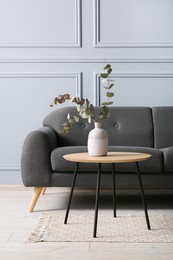 Photo of Stylish sofa and eucalyptus branches in vase on coffee table near grey wall