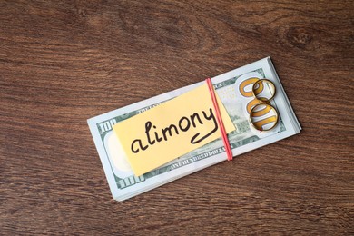 Card with word Alimony, dollar banknotes and rings on wooden table, top view