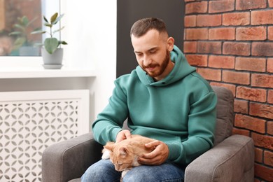 Photo of Man petting cute ginger cat on armchair at home