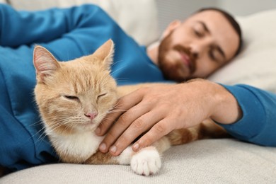 Man petting cute ginger cat on sofa at home, selective focus