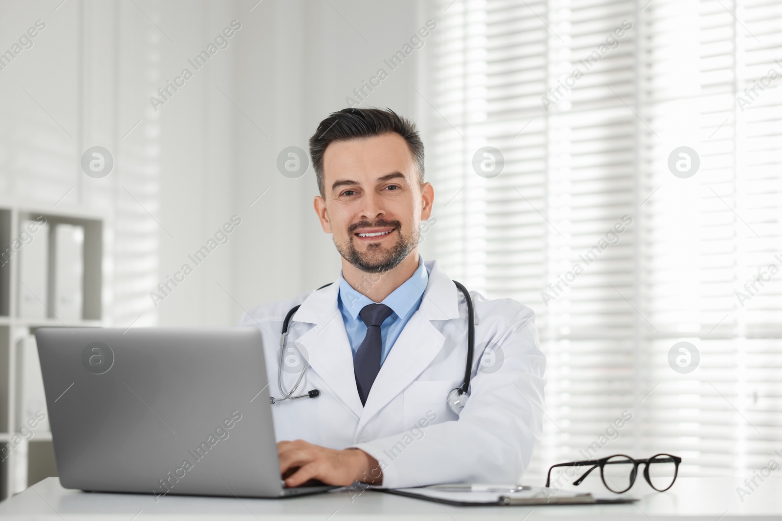 Photo of Smiling doctor working with laptop at table in clinic