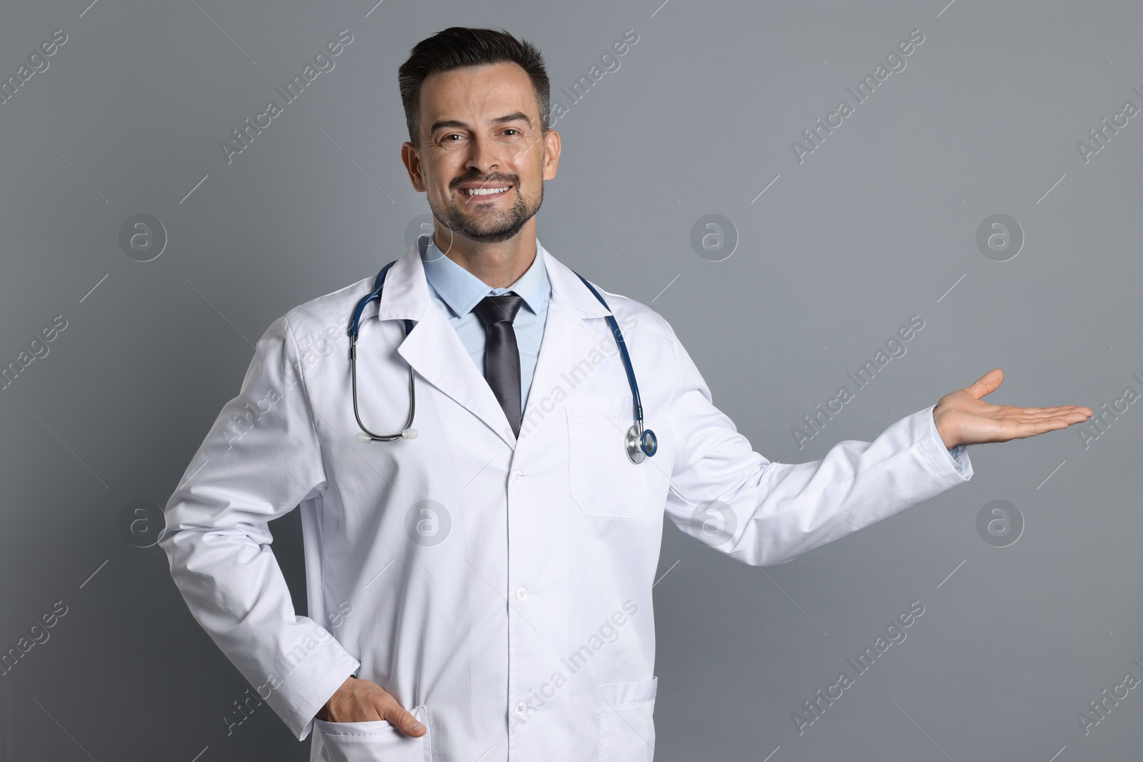 Photo of Smiling doctor with stethoscope pointing at something on grey background