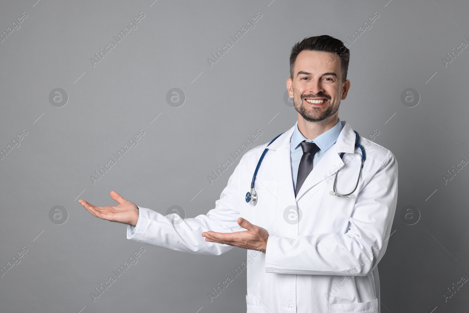 Photo of Smiling doctor with stethoscope pointing at something on grey background