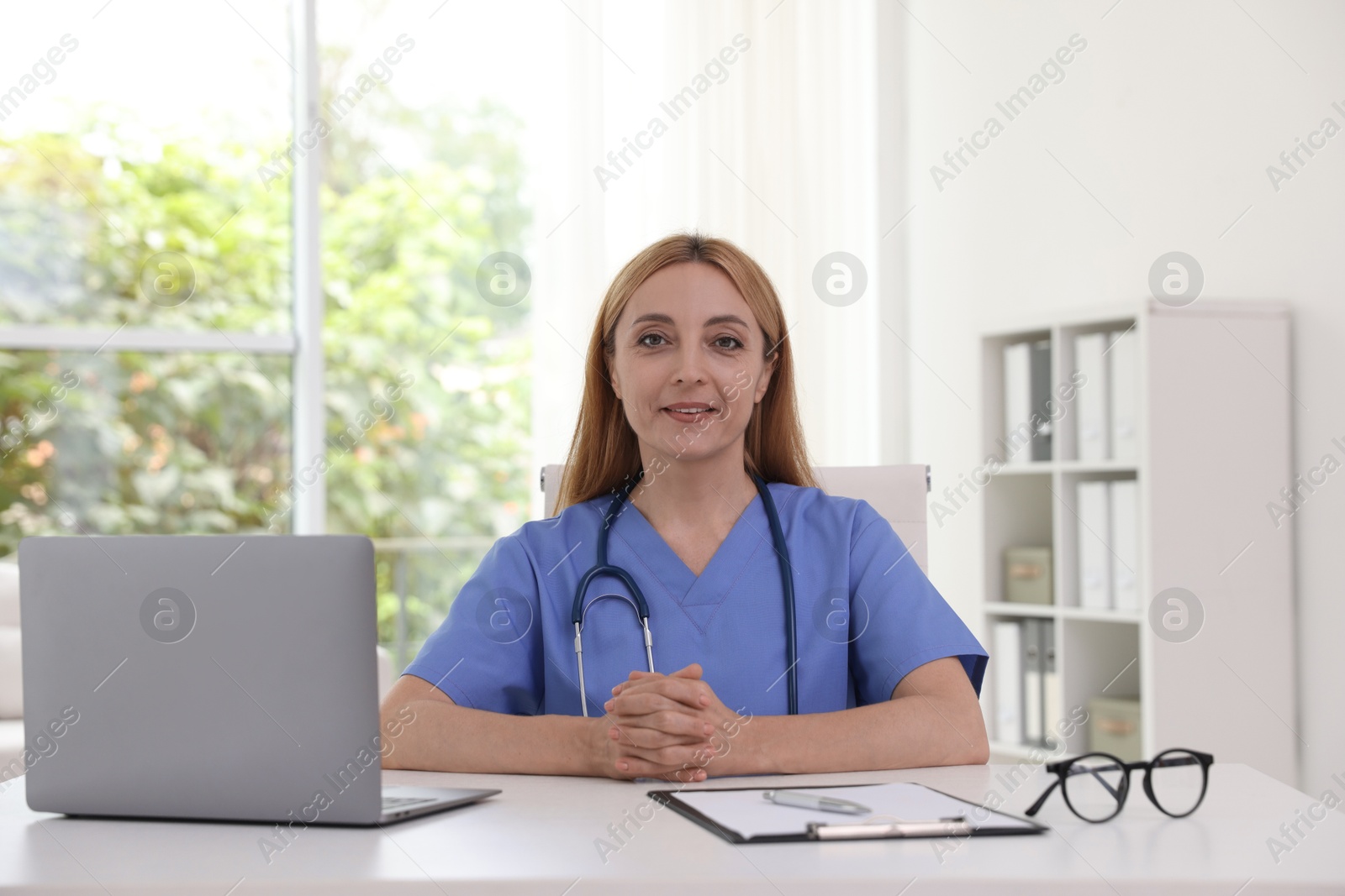 Photo of Doctor with stethoscope, laptop and clipboard at table in clinic