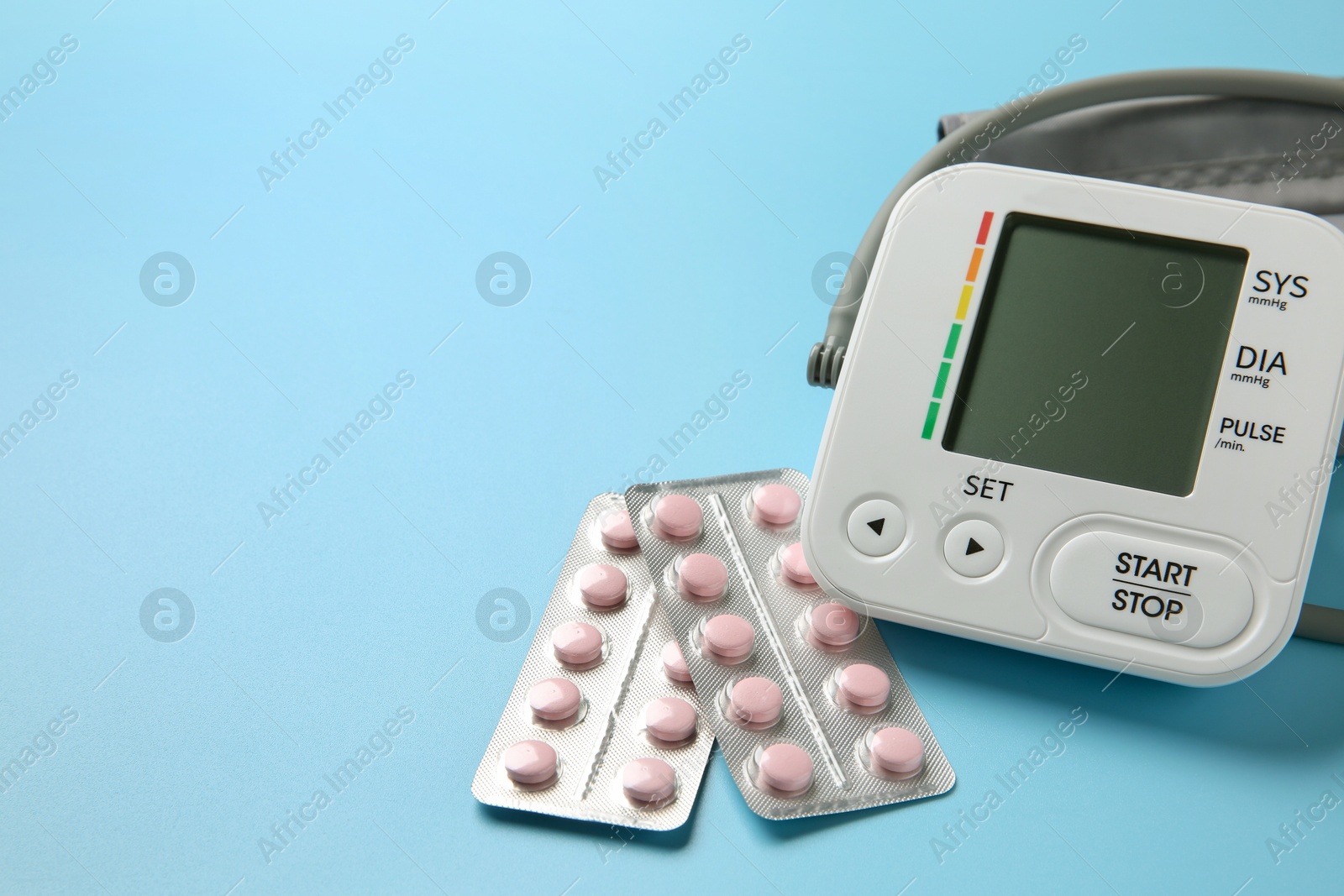 Photo of Blood pressure measuring device and pills on light blue background, closeup. Space for text