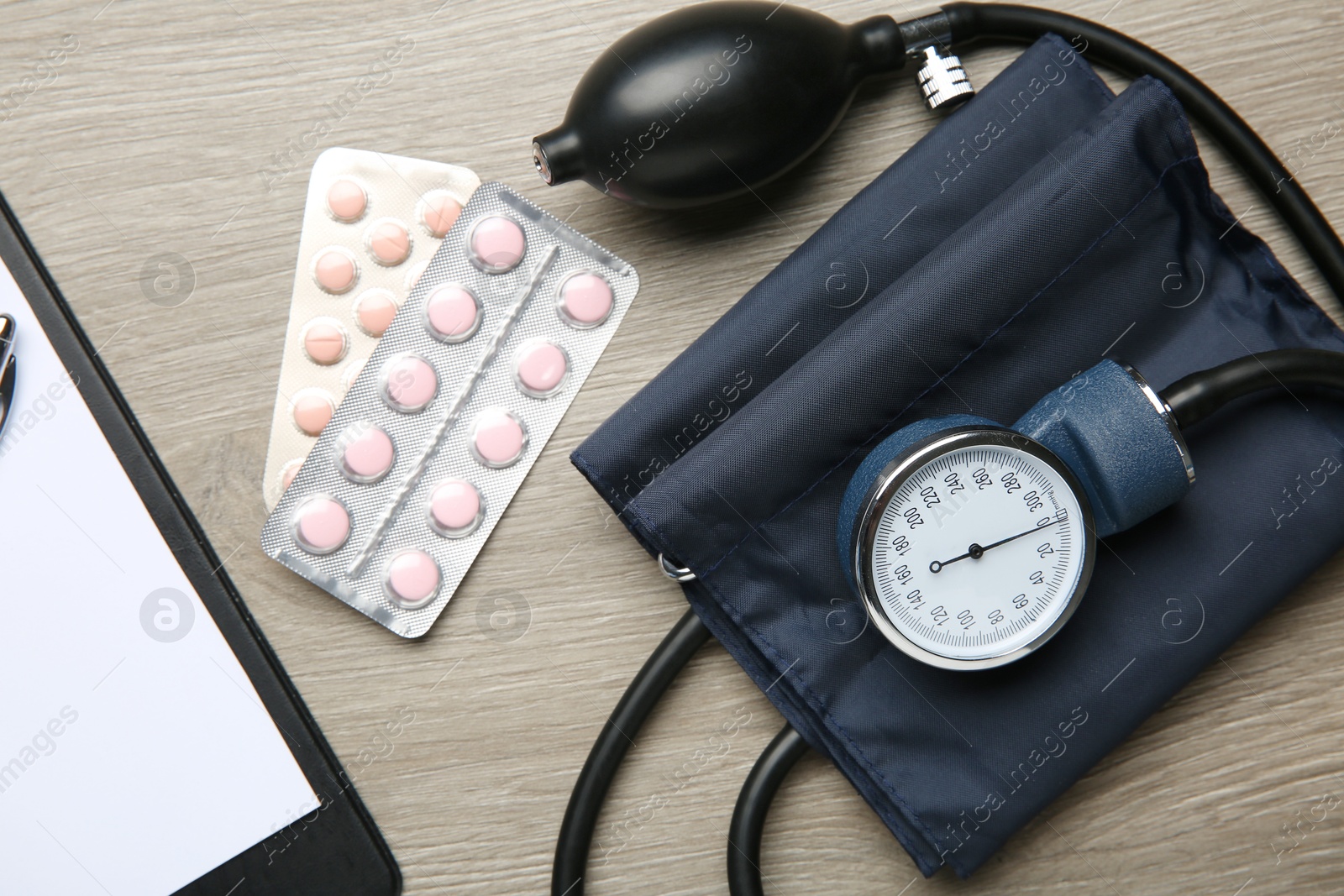 Photo of Blood pressure measuring device, pills and clipboard on wooden table, top view