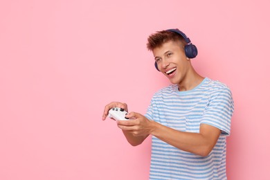 Happy young man in headphones playing video games with controller on pink background, space for text