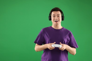 Happy young man in headphones playing video games with controller on green background, space for text