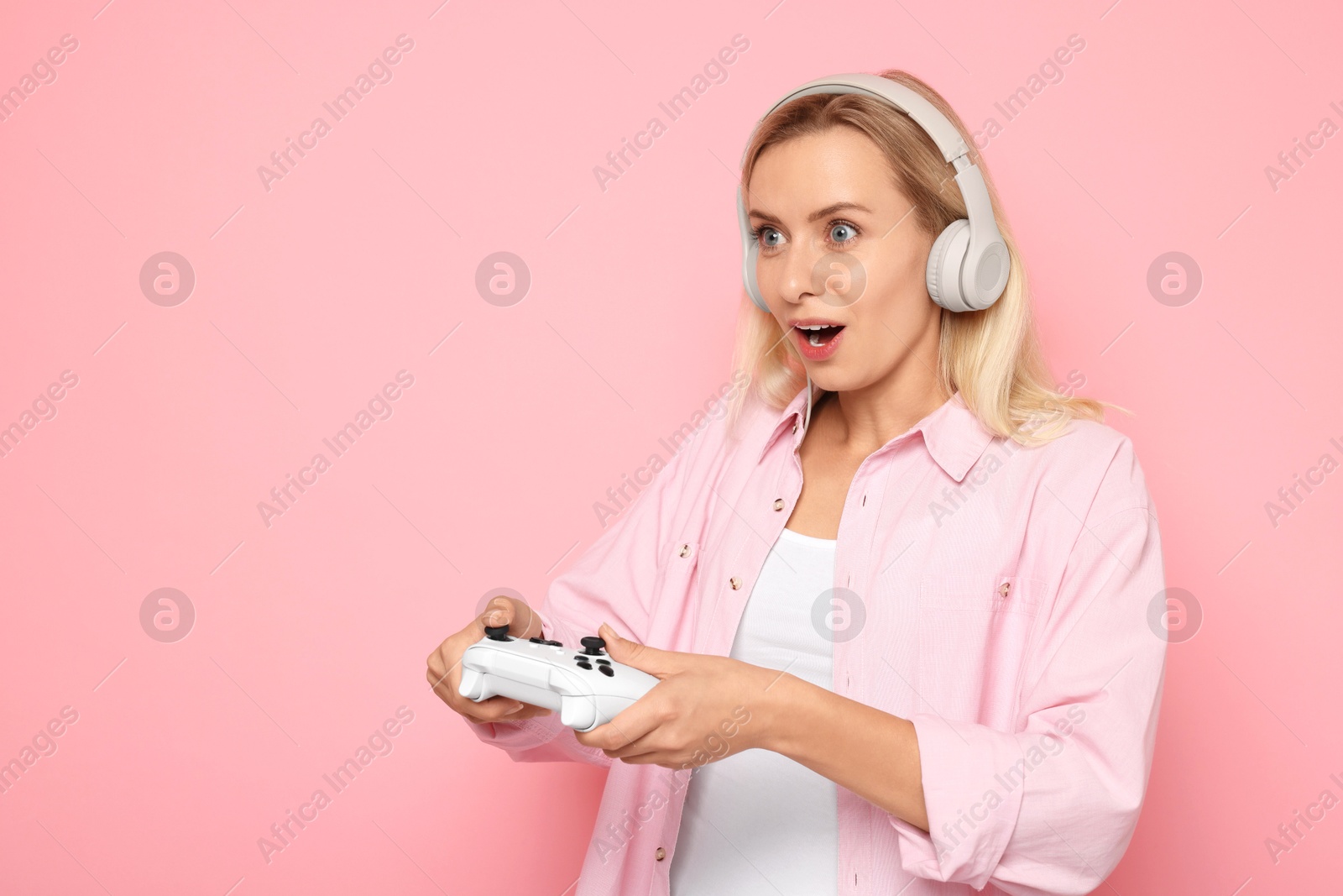 Photo of Surprised woman in headphones playing video games with controller on pink background, space for text