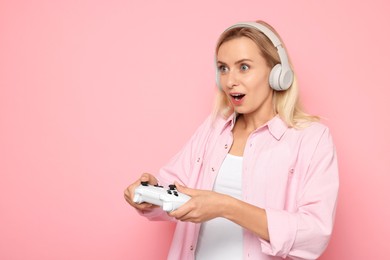 Photo of Surprised woman in headphones playing video games with controller on pink background, space for text