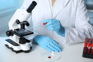 Photo of Laboratory testing. Doctor dripping blood sample onto glass slide while working with microscope at table indoors, closeup