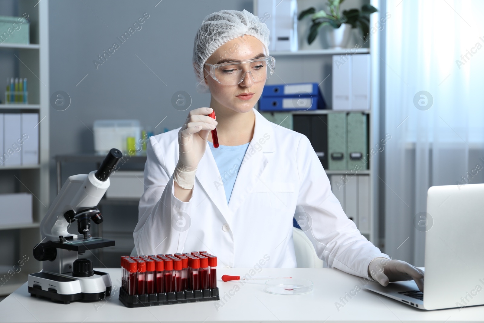 Photo of Laboratory testing. Doctor holding test tube with blood sample while working on laptop at table indoors