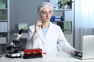 Photo of Laboratory testing. Doctor holding test tube with blood sample while working on laptop at table indoors