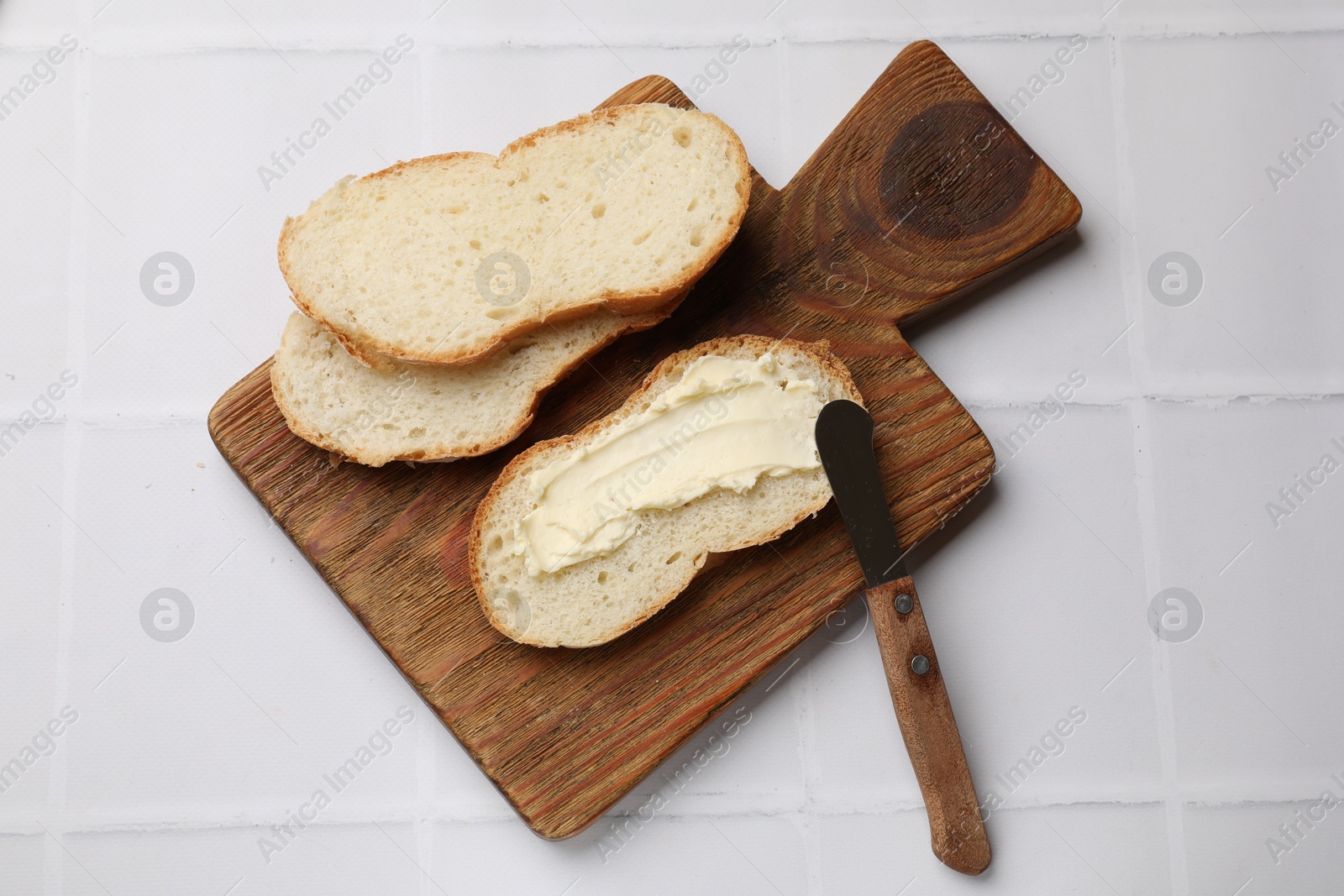 Photo of Fresh bread with butter and knife on white tiled table, top view