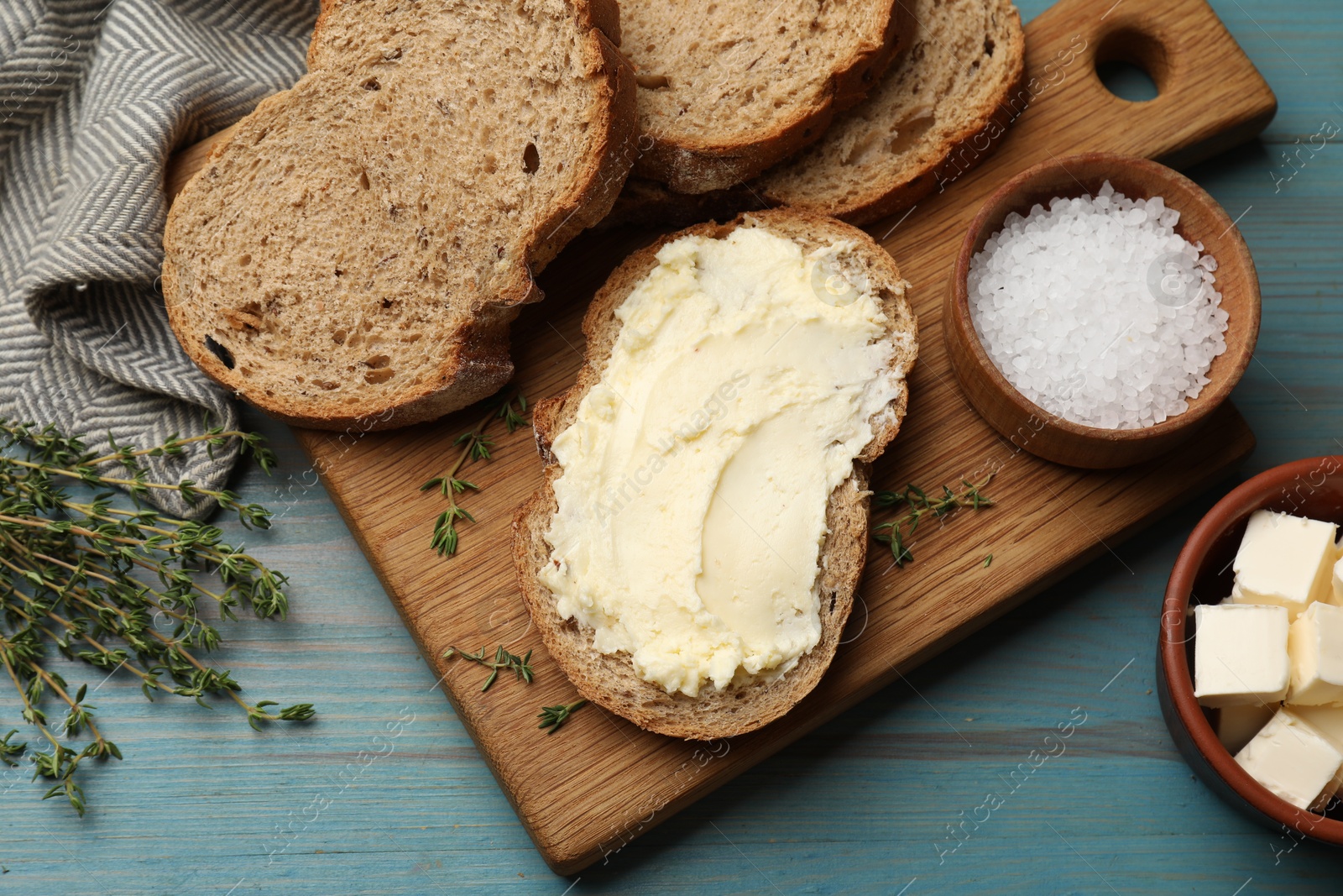 Photo of Fresh bread with butter, salt and thyme on blue wooden table, flat lay