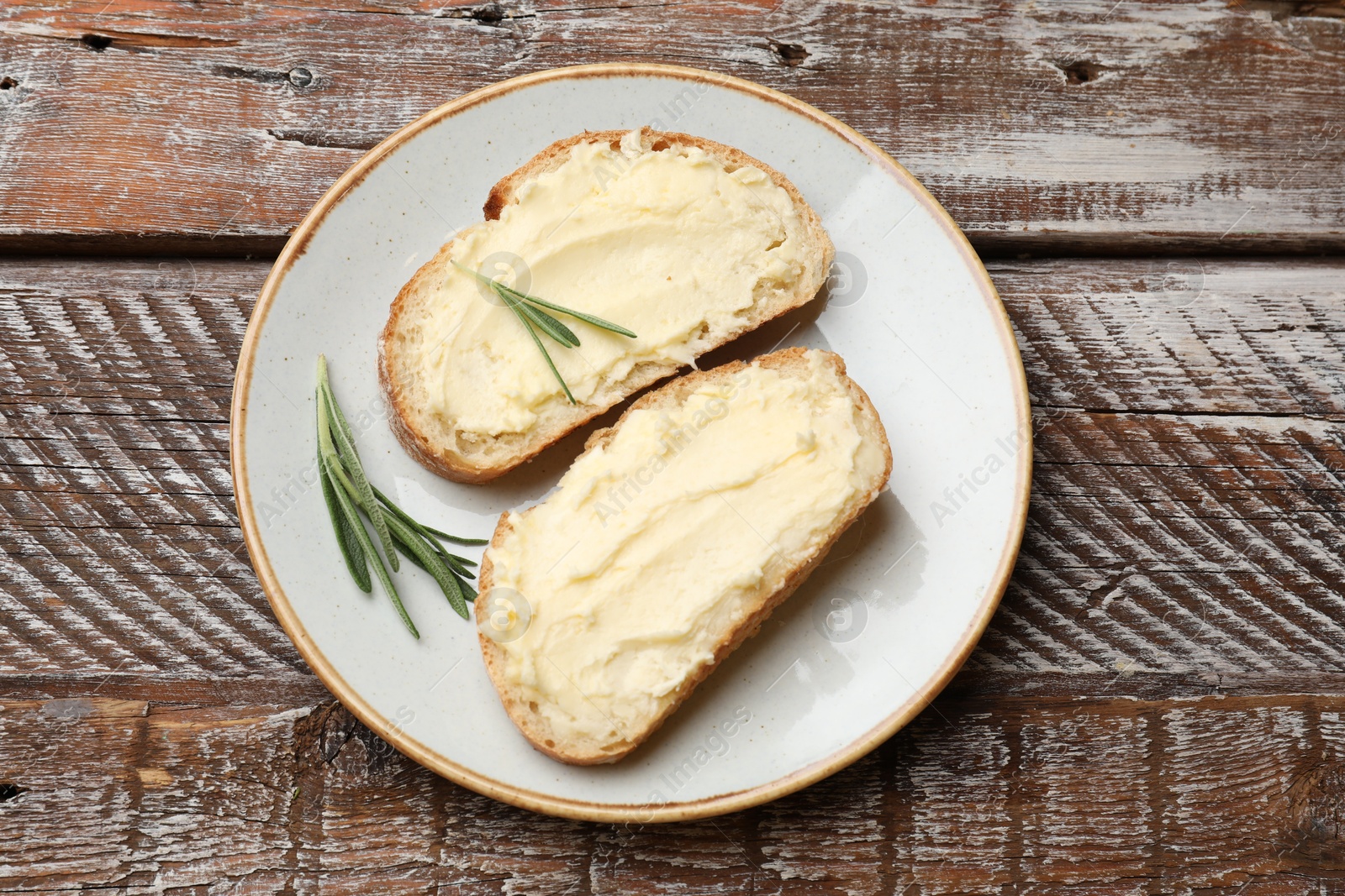 Photo of Fresh bread with butter and rosemary on wooden table, top view