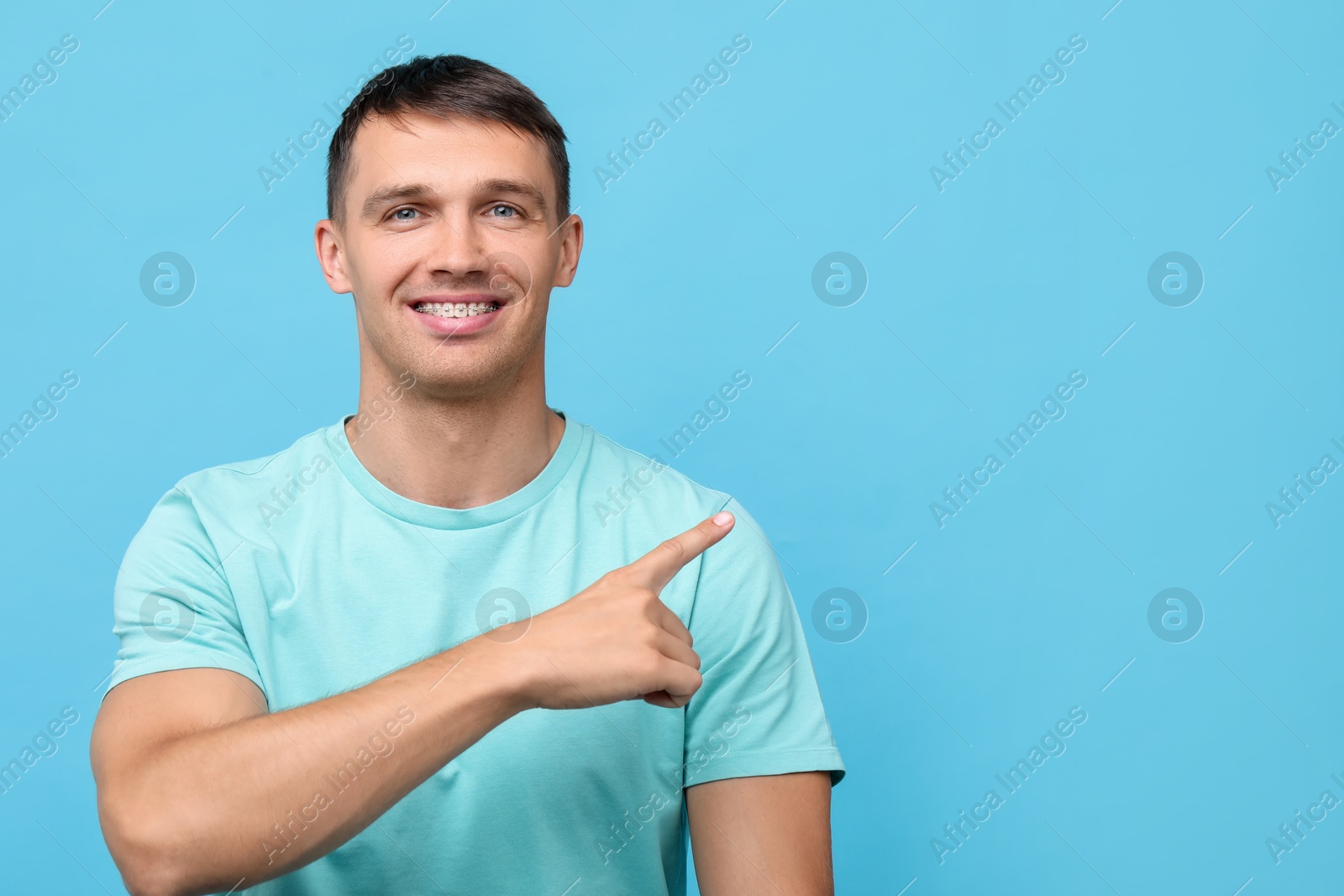 Photo of Smiling man with dental braces pointing at something on light blue background. Space for text