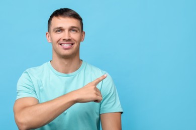 Photo of Smiling man with dental braces pointing at something on light blue background. Space for text