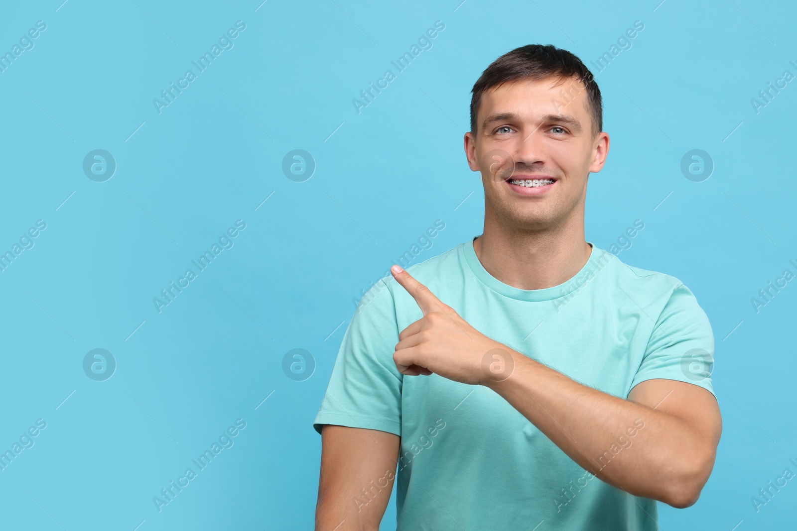 Photo of Smiling man with dental braces pointing at something on light blue background. Space for text