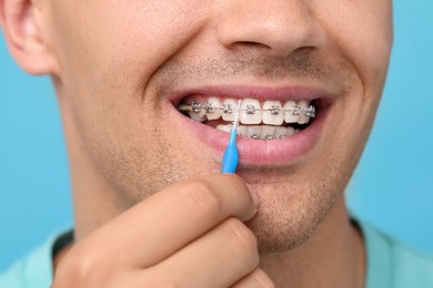 Photo of Man with dental braces cleaning teeth using interdental brush on light blue background, closeup