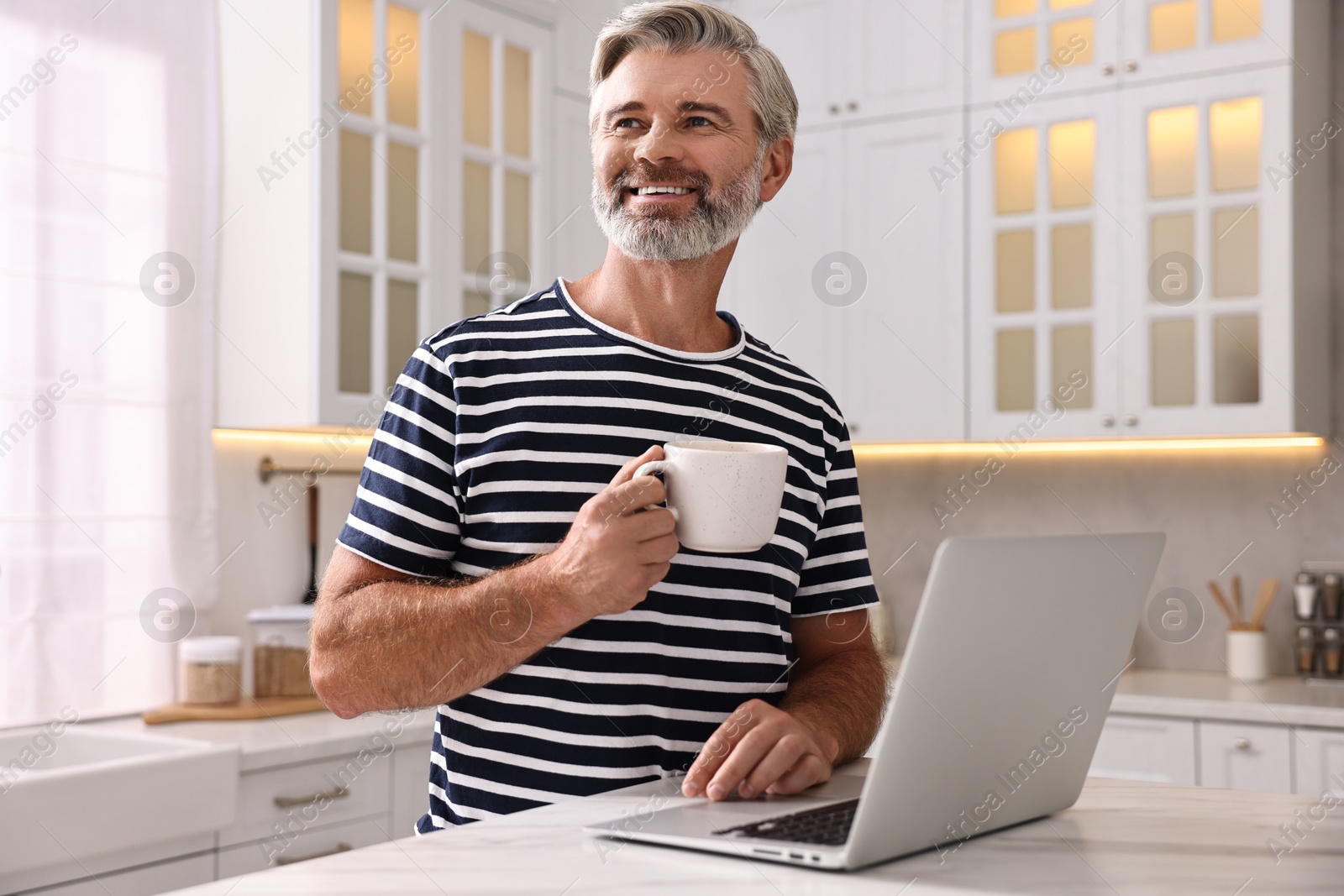 Photo of Happy middle aged man with cup of drink and laptop at white marble table in kitchen