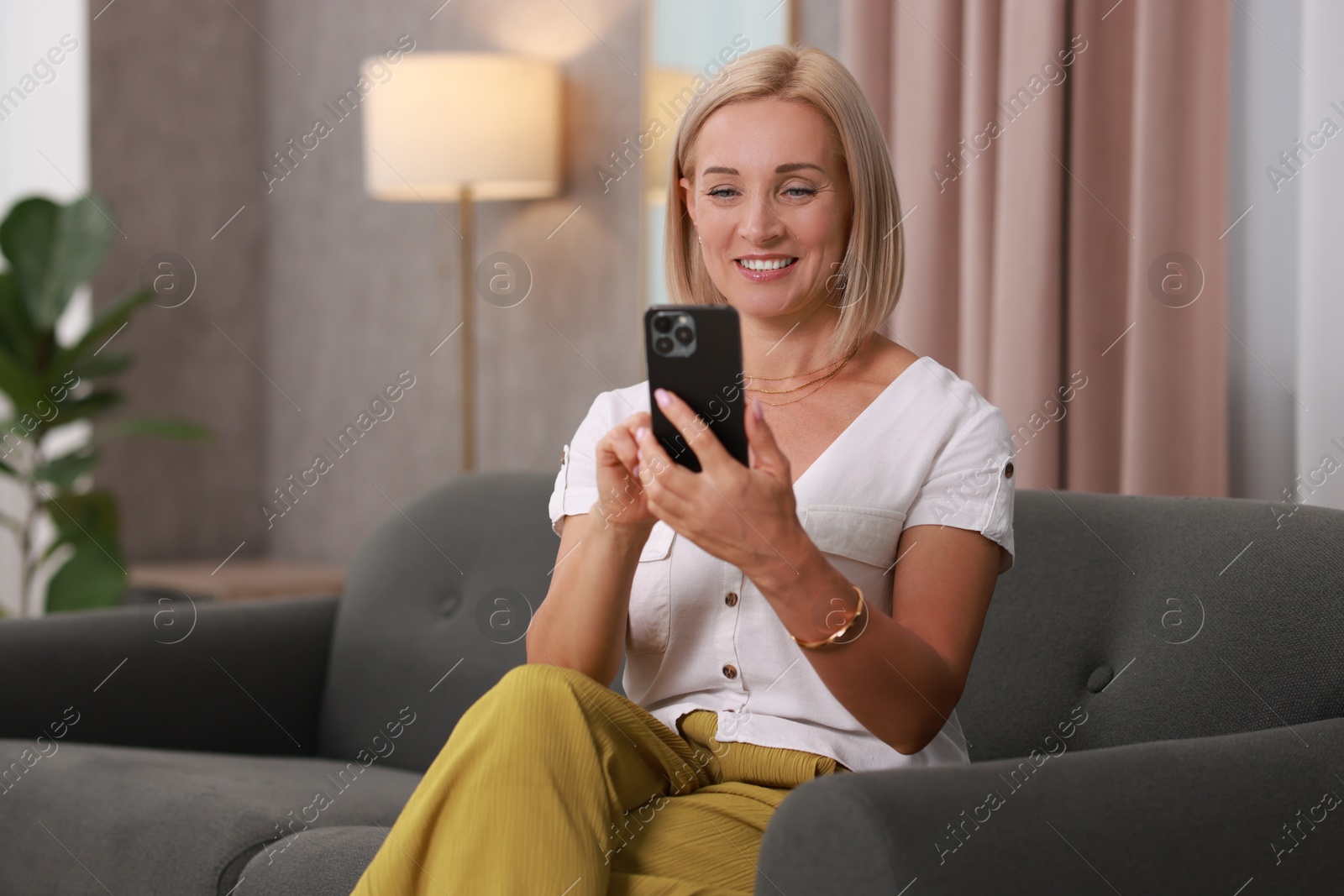 Photo of Portrait of smiling middle aged woman with smartphone on sofa at home