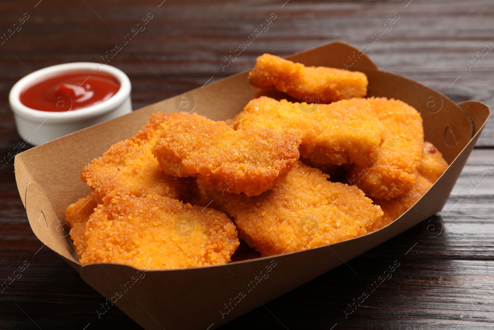 Photo of Tasty chicken nuggets in paper container and sauce on wooden table, closeup