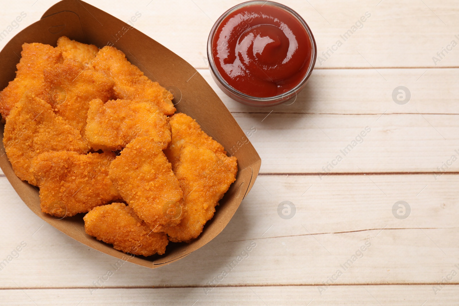 Photo of Tasty chicken nuggets in paper container and sauce on wooden table, top view. Space for text