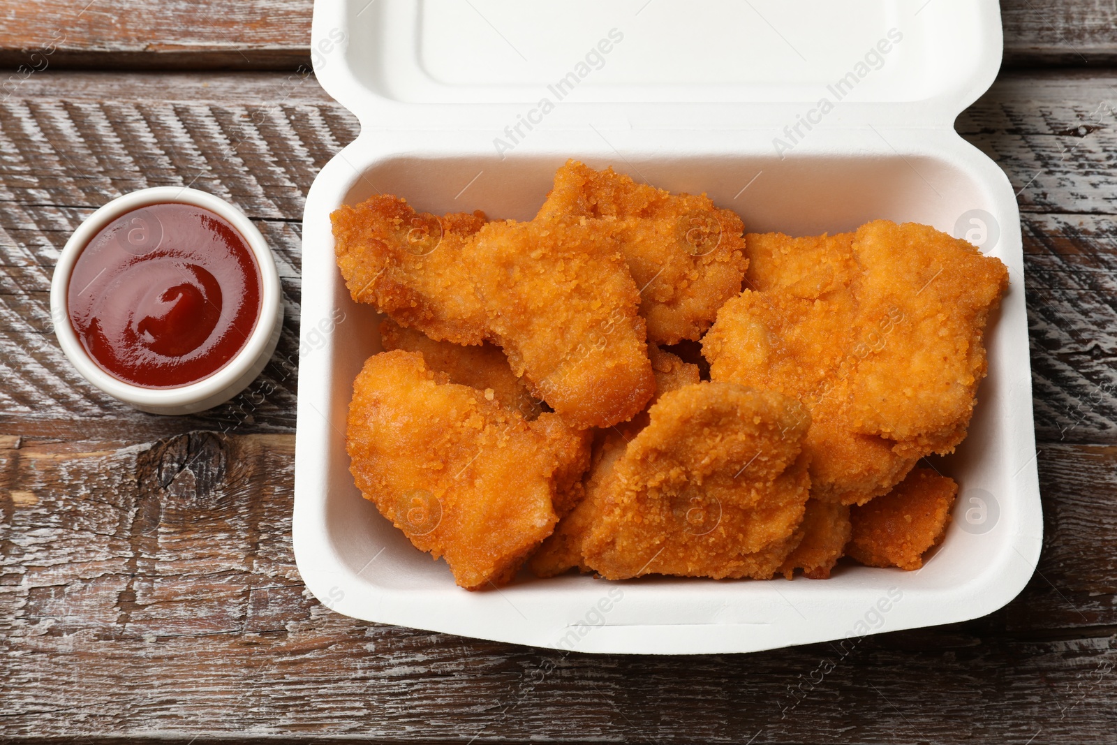 Photo of Delicious chicken nuggets in takeaway container and ketchup on wooden table, above view