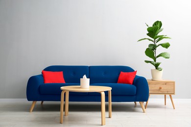 Image of Comfortable blue sofa, tables and houseplant near grey wall in room