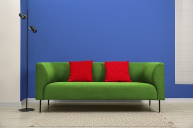 Image of Stylish green sofa with red pillows and lamp near blue wall indoors