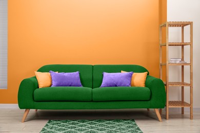 Image of Stylish green sofa with pillows and shelving unit near orange wall indoors