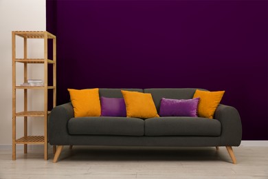 Image of Stylish grey sofa with pillows and shelving unit near purple wall indoors