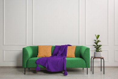 Image of Stylish green sofa with cushions, blanket and table near white wall