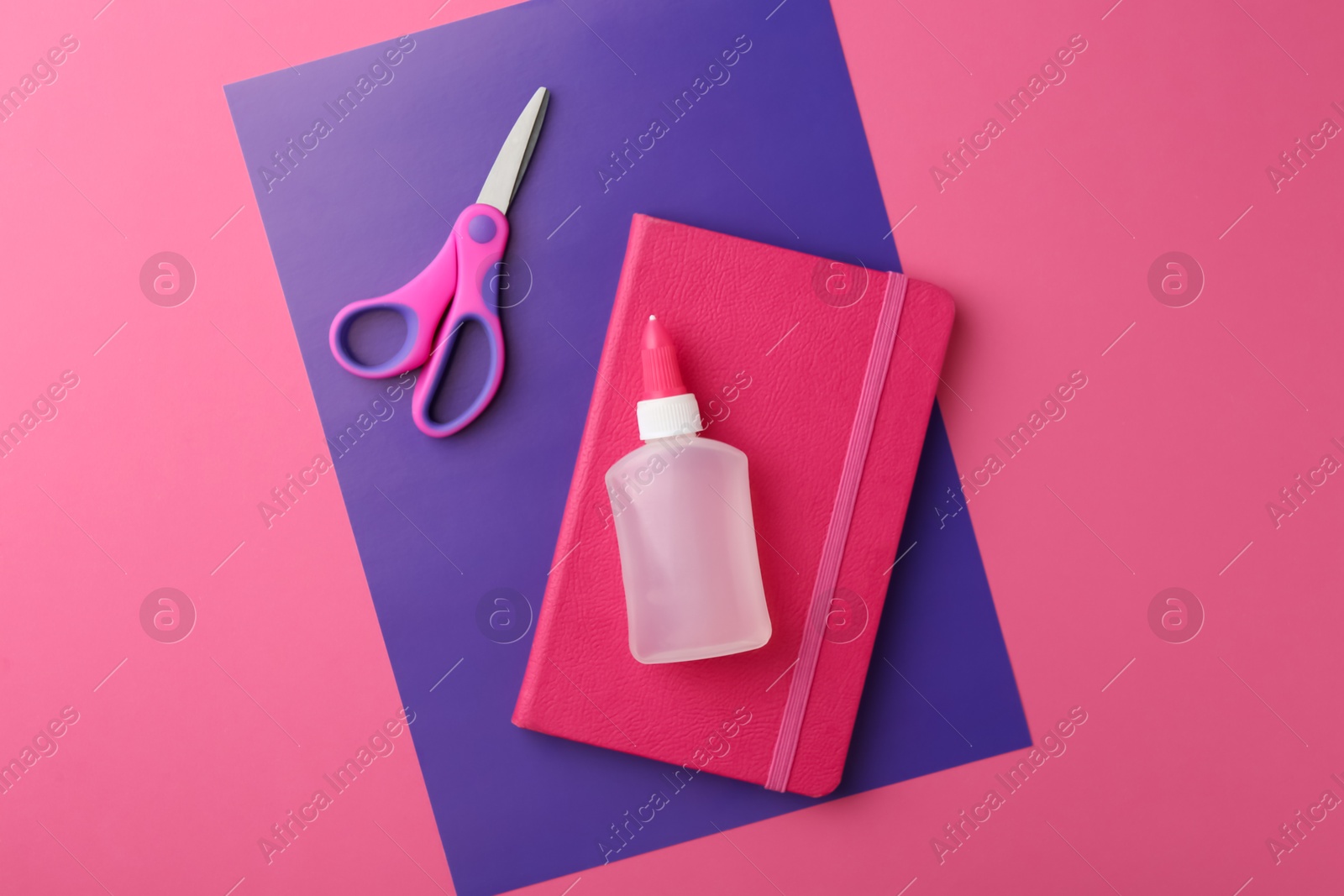 Photo of Glue, notebook, purple paper and scissors on pink background, top view