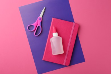 Glue, notebook, purple paper and scissors on pink background, top view