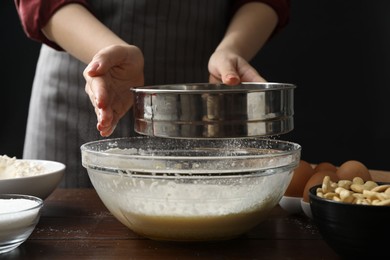 Photo of Making cashew cookies. Woman sieving flour into dough at wooden table, closeup