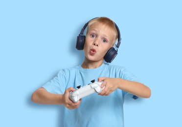 Cute little boy in headphones playing video game with controller on light blue background