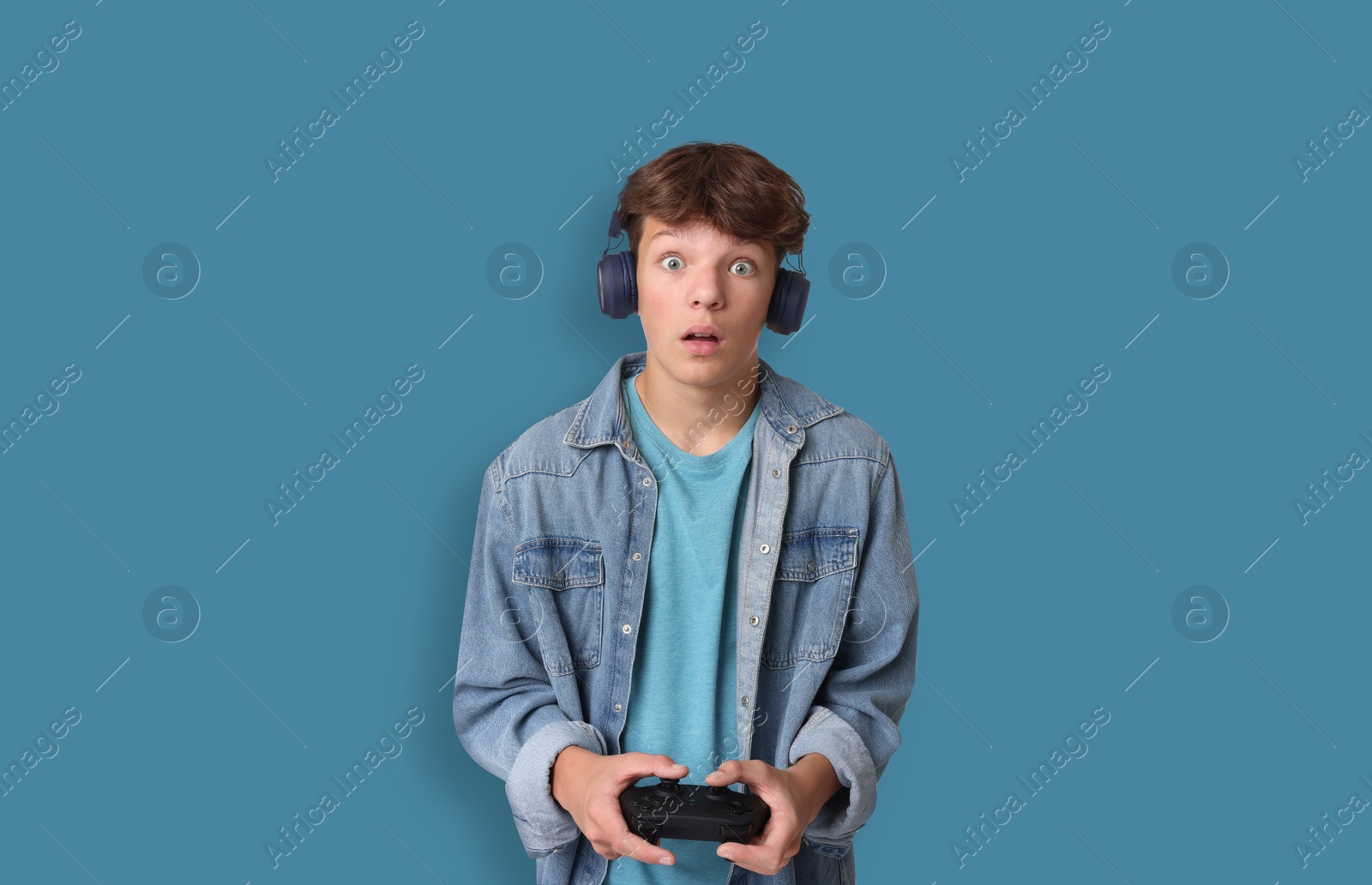 Image of Emotional teenage boy in headphones playing video game with controller on light blue background