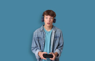 Emotional teenage boy in headphones playing video game with controller on light blue background