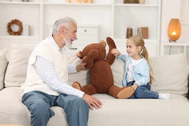 Grandpa and his granddaughter playing with toy bear on sofa at home