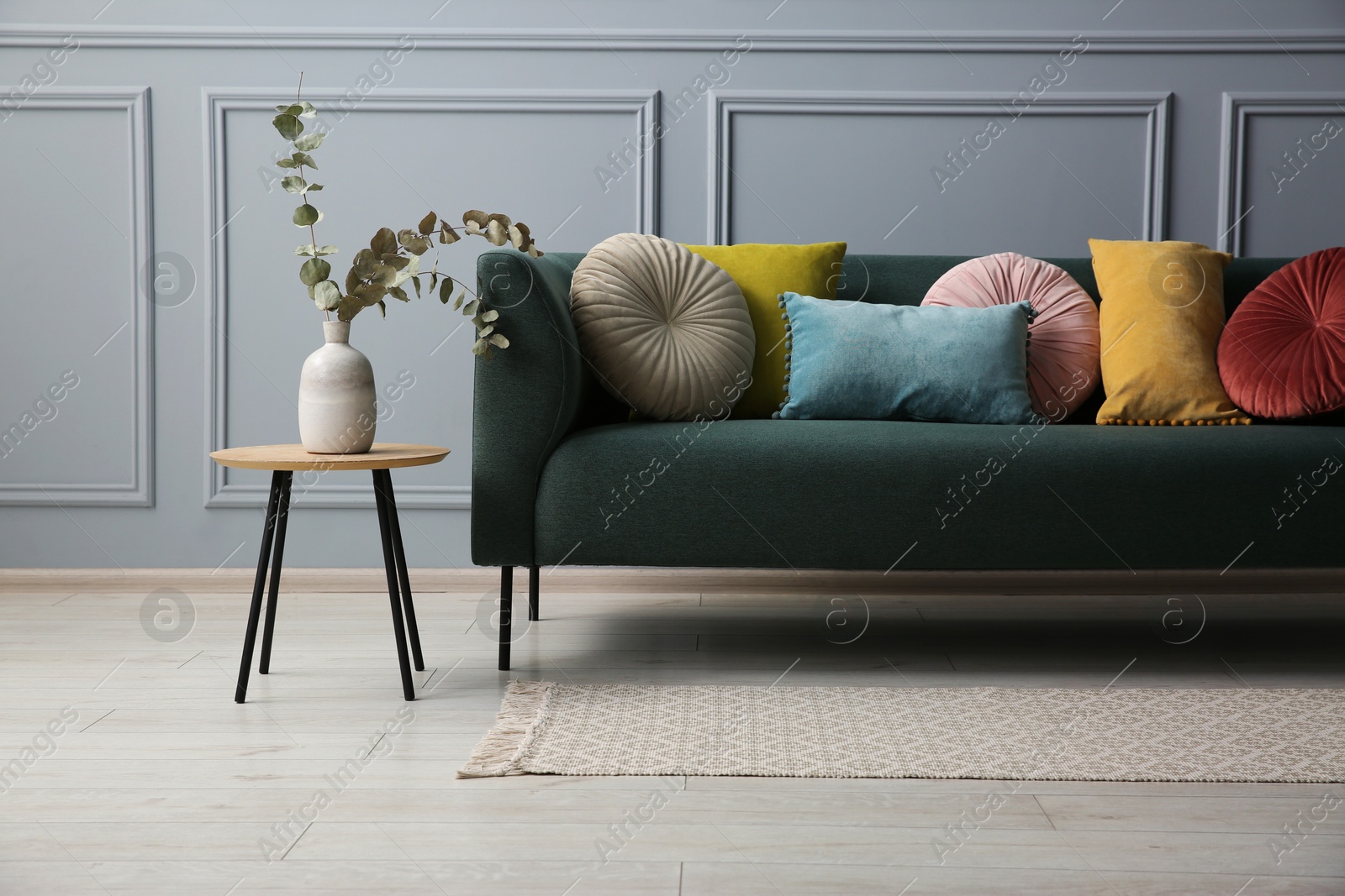 Photo of Stylish sofa with cushions and eucalyptus branches in vase on side table near grey wall