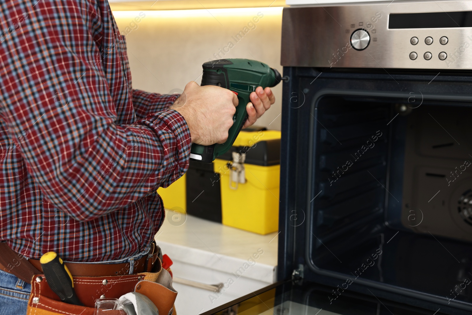 Photo of Repairman with electric screwdriver fixing oven indoors, closeup