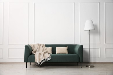 Photo of Stylish sofa with cushions, blanket and floor lamp near white wall